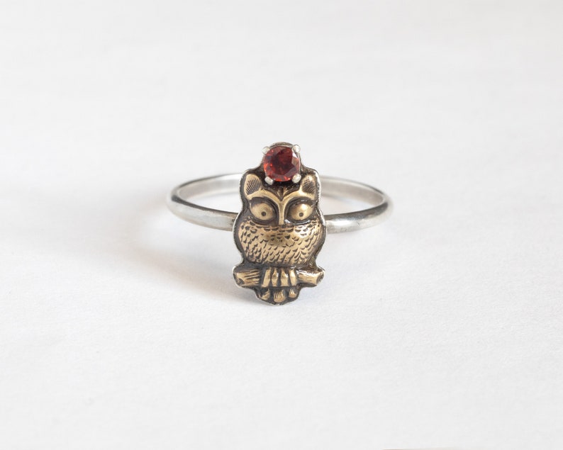 Tiny brass owl ring with a garnet above the head and a thin silver band. white background.