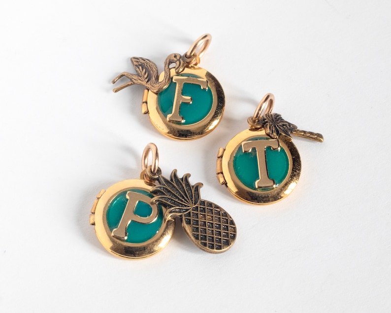 Group of small gold round lockets, with initials set in teal blue resin. Each has a tropical-themed brass charm- flamingo, palm tree, and pineapple. Flat on a white background.