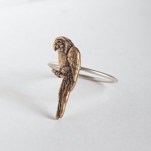 brass parrot charm on a thin silver band. at an angle on a white surface