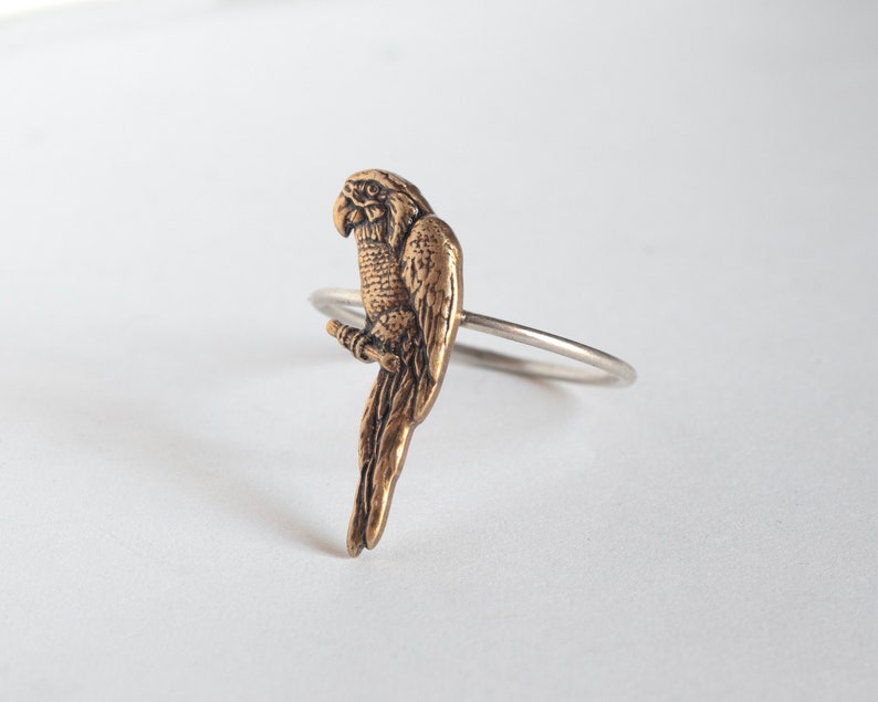 brass parrot charm on a thin silver band. at an angle on a white surface