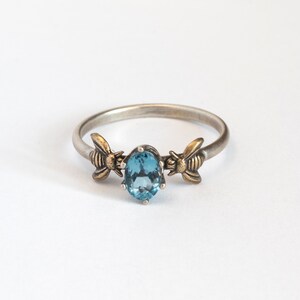 London blue oval gemstone ring, in a prong setting with a brass bee on each side. Shown on a white background.