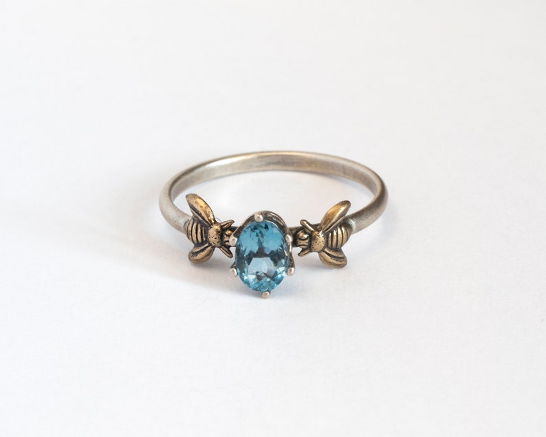 London blue oval gemstone ring, in a prong setting with a brass bee on each side. Shown on a white background.