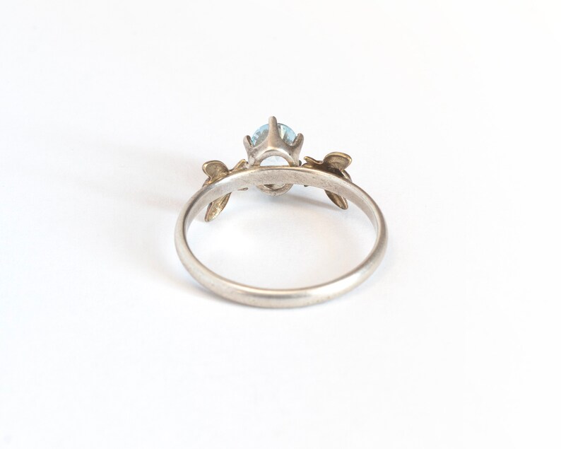 Sky blue oval gemstone ring, in a prong setting with a brass bee on each side. Shown from the back on a white background.