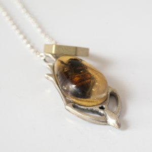 Real honeybee preserved in resin with gold backing in a teardrop shape. Set into a hand carved silver pendant in an art deco style. On a chain with a brass hexagon, shown at an angle on a white background.