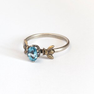 London blue oval gemstone ring, in a prong setting with a brass bee on each side. Shown at an angle on a white background.