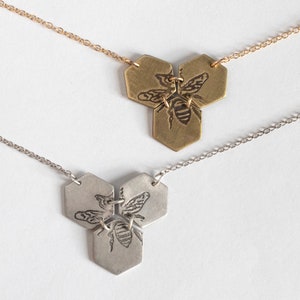 three hexagon pendants with etched bees, one in brass and one in silver, on a white background