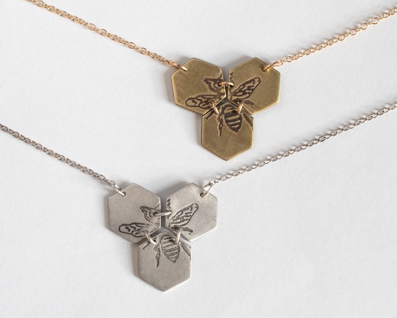 three hexagon pendants with etched bees, one in brass and one in silver, on a white background