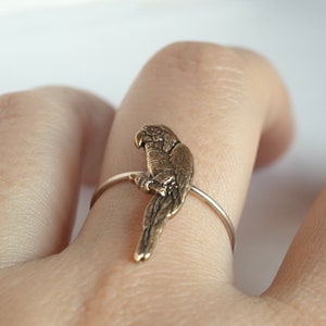 brass parrot charm on a thin silver band. Worn on a hand on a white surface