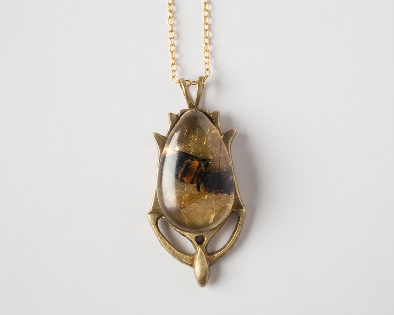 Real honeybee preserved in resin with gold backing in a teardrop shape. Set into a hand carved brass pendant in an art deco style. Shown flat on a white background.