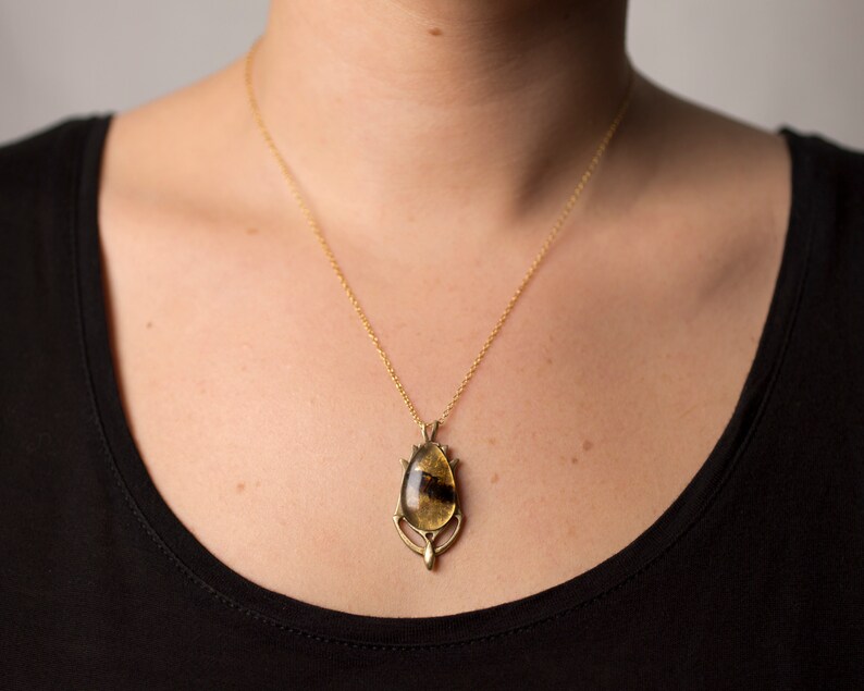 Real honeybee preserved in resin with gold backing in a teardrop shape. Set into a hand carved brass pendant in an art deco style. Worn by a model wearing a black tee with white background.