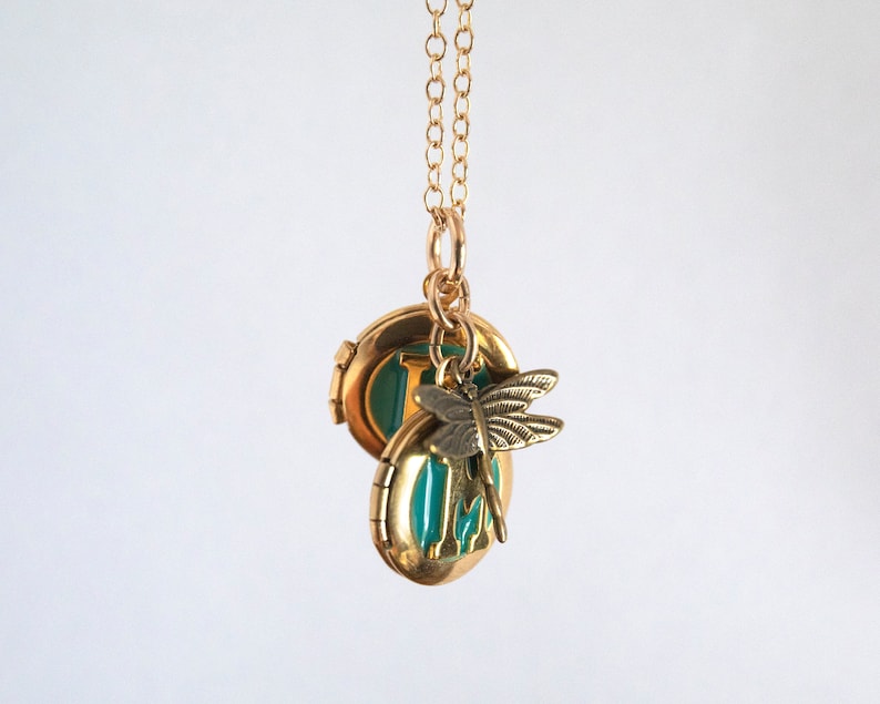 Two small gold round lockets with initials set in teal blue resin, with a brass dragonfly charm. All together on a cable chain with a white background.