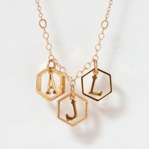 a gold necklace with 3 initials in hexagons in a row, hanging in front of a white background