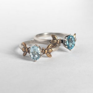 London blue and sky blue topaz oval gemstone rings, in a prong setting with a brass bee on each side. Shown laying on a white background.
