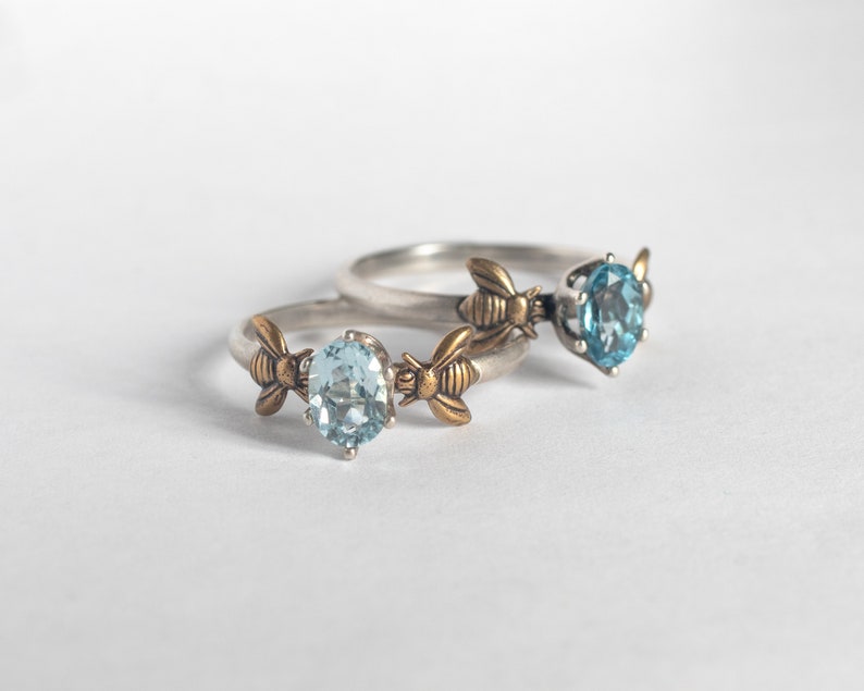 London blue and sky blue topaz oval gemstone rings, in a prong setting with a brass bee on each side. Shown laying on a white background.