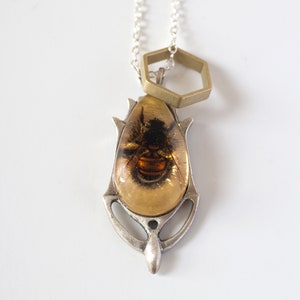 Real honeybee preserved in resin with gold backing in a teardrop shape. Set into a hand carved silver pendant in an art deco style. On a chain with a brass hexagon, flat on a white background.