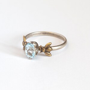 Sky blue oval gemstone ring, in a prong setting with a brass bee on each side. Shown at an angle on a white background.