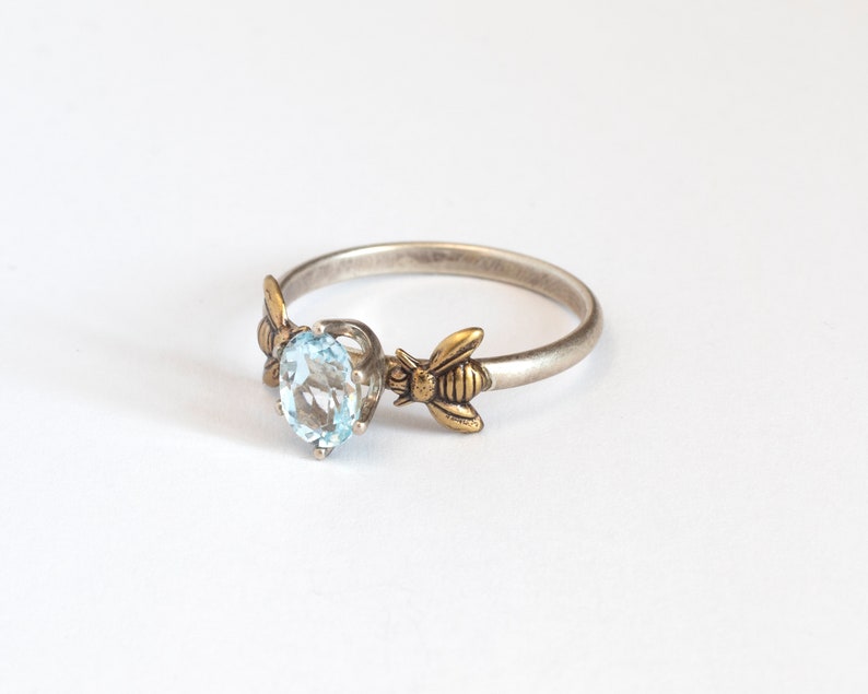 Sky blue oval gemstone ring, in a prong setting with a brass bee on each side. Shown at an angle on a white background.