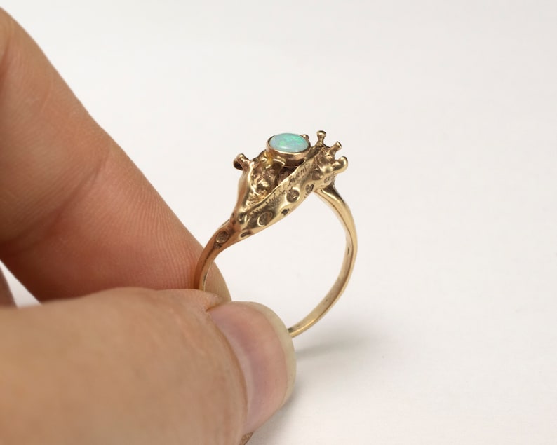 14k gold double giraffe head ring, with opal cabochon, held by fingers on white background