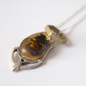 Real honeybee preserved in resin with gold backing in a teardrop shape. Set into a hand carved silver pendant in an art deco style. On a chain with a brass hexagon, shown at an angle on a white background.