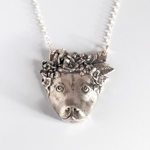 Puede incluir: Un collar de plata con un colgante que representa la cabeza de un perro con una corona de flores. La cabeza del perro está esculpida con detalle y las flores son delicadas y realistas.