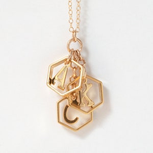 a gold necklace with 3 initials in hexagons in a group, hanging in front of a white background