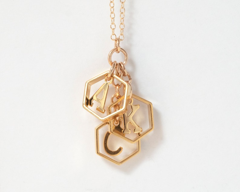 a gold necklace with 3 initials in hexagons in a group, hanging in front of a white background