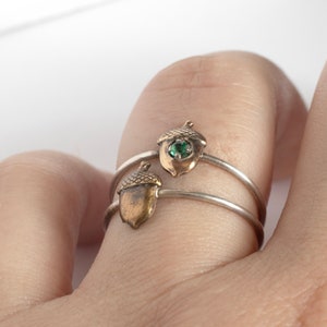 May include: Two silver rings with acorn-shaped charms. The charms are gold-toned and have a green gemstone in the center.