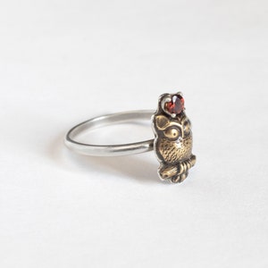 Tiny brass owl ring with garnet above the head and a thin silver band. At an angle on a white background.