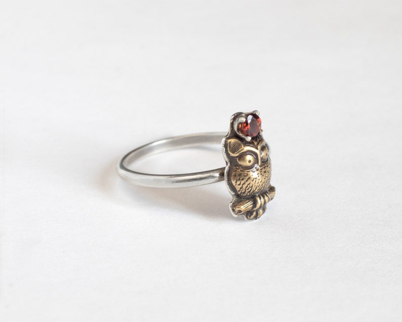 Tiny brass owl ring with garnet above the head and a thin silver band. At an angle on a white background.