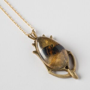 Real honeybee preserved in resin with gold backing in a teardrop shape. Set into a hand carved brass pendant in an art deco style. Shown at an angle on a white background.