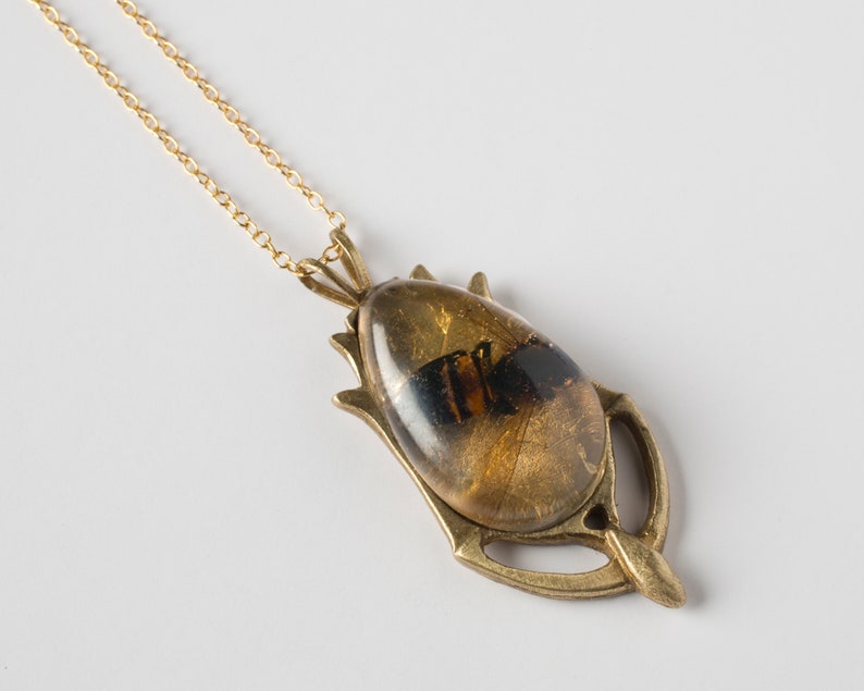 Real honeybee preserved in resin with gold backing in a teardrop shape. Set into a hand carved brass pendant in an art deco style. Shown at an angle on a white background.