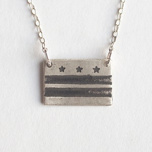 May include: A silver necklace with a rectangular pendant featuring a black and white design of the Washington D.C. flag.
