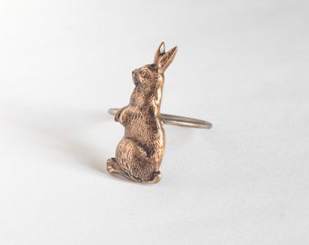 May include: A gold-toned metal rabbit napkin ring. The rabbit is standing on its hind legs with its front paws raised. The ring is open and ready to hold a napkin.