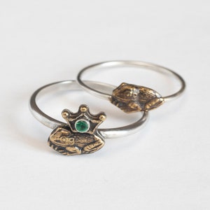Puede incluir: Dos anillos de plata con dijes de rana. Una rana lleva una corona con una piedra preciosa verde.