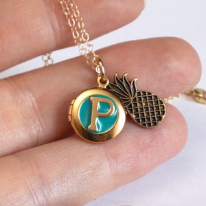 Small gold round locket, with initial P set in teal blue resin, with a brass pineapple charm. Held in a hand with a white background.