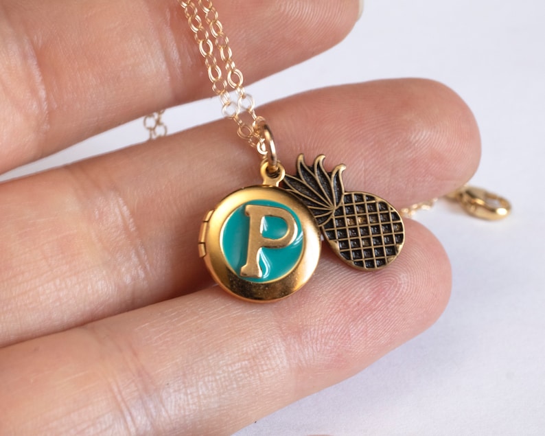 Small gold round locket, with initial P set in teal blue resin, with a brass pineapple charm. Held in a hand with a white background.