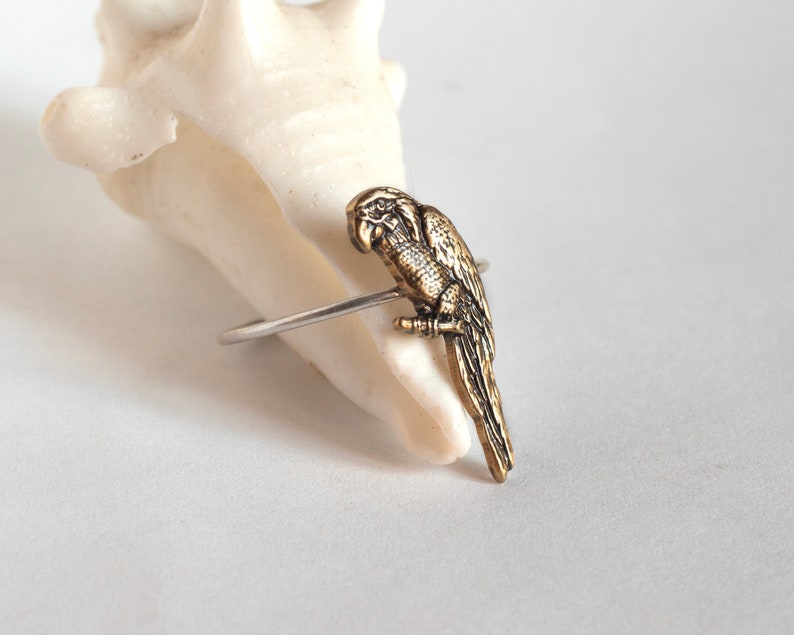 brass parrot charm on a thin silver band. STyled on a seashell at an angle on a white surface