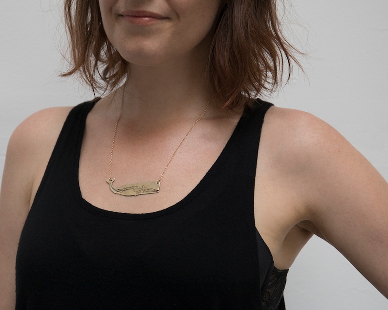 brass whale necklace with etched skeleton, worn by a model wearing a black tank top in front of a white wall