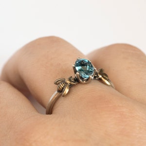 London blue oval gemstone ring, in a prong setting with a brass bee on each side. Shown on a hand on a white background.