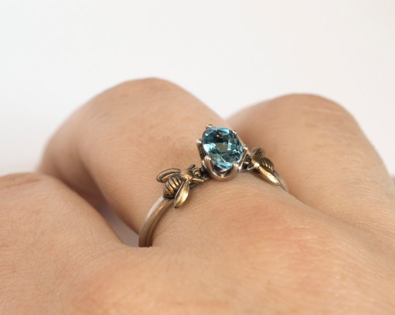 London blue oval gemstone ring, in a prong setting with a brass bee on each side. Shown on a hand on a white background.