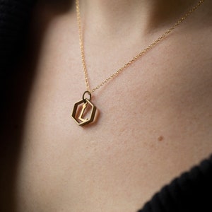 close up of model wearing a gold hexagon pendant with an initial inside and a black v-neck sweater.