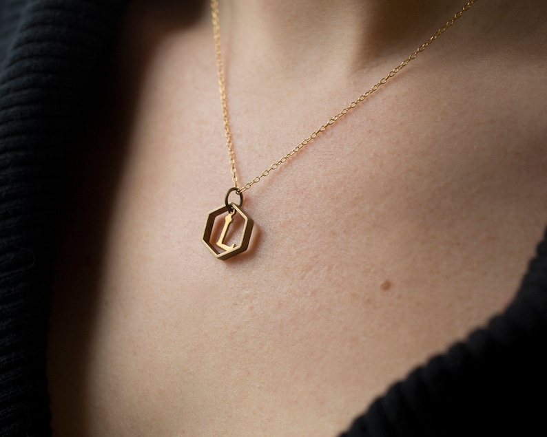 close up of model wearing a gold hexagon pendant with an initial inside and a black v-neck sweater.