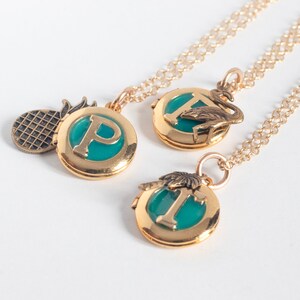 Group of small gold round lockets, with initials set in teal blue resin. Each has a tropical-themed brass charm- flamingo, palm tree, and pineapple. Shown at an angle on a white background.