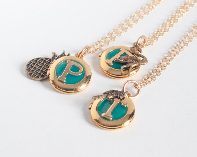 Group of small gold round lockets, with initials set in teal blue resin. Each has a tropical-themed brass charm- flamingo, palm tree, and pineapple. Shown at an angle on a white background.