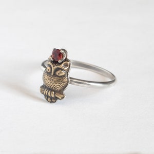 Tiny brass owl ring with garnet above the head and a thin silver band. At an angle on a white background.