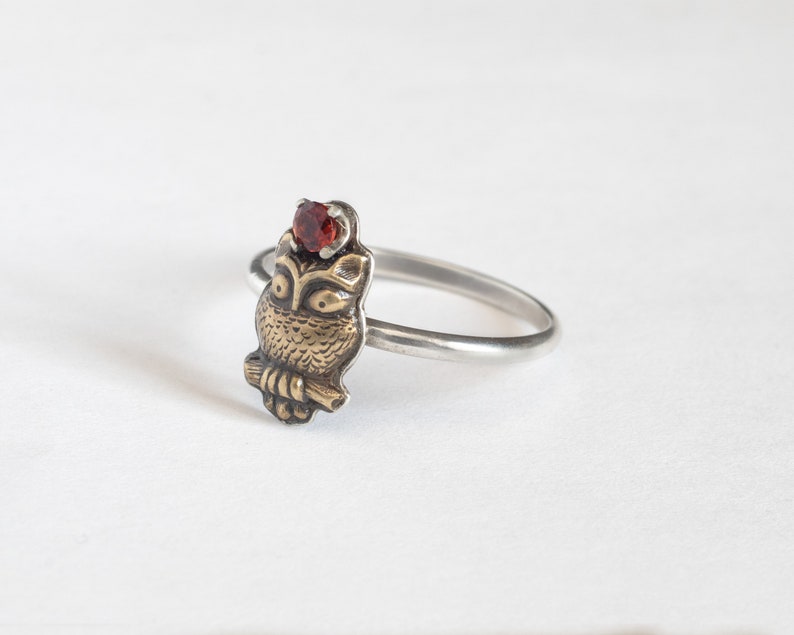 Tiny brass owl ring with garnet above the head and a thin silver band. At an angle on a white background.