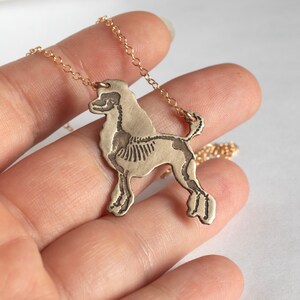 brass poodle silhouette necklace with etched skeleton, held by a hand on a white background