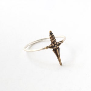 Puede incluir: Un anillo plateado con un diseño de daga en color bronce. La daga tiene una empuñadura detallada y una hoja puntiaguda. El anillo se presenta sobre un fondo blanco, resaltando el diseño de la joya.