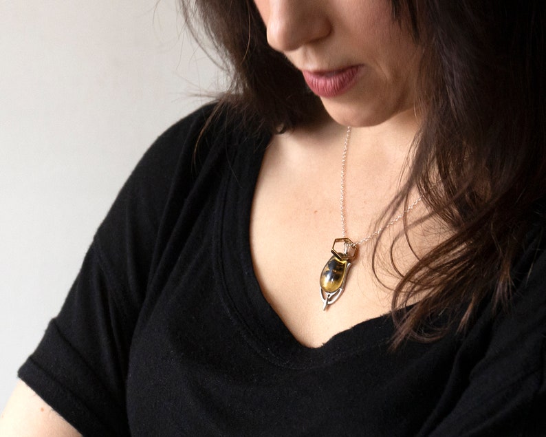 Real honeybee preserved in resin with gold backing in a teardrop shape. Set into a hand carved silver pendant in an art deco style. Worn by a model wearing a black tee with white background.