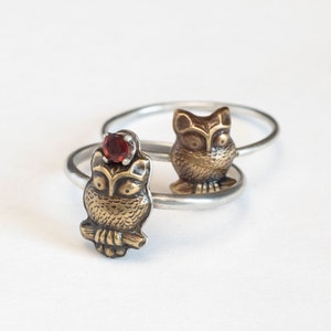 two brass owl rings on silver bands, one with a garnet and one without. white background.